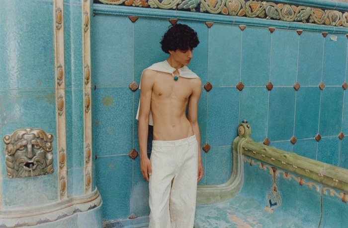 Model Yoesry Detre standing shirtless in white trousers and neck wrap against ornate turquoise ceramic tile walls.