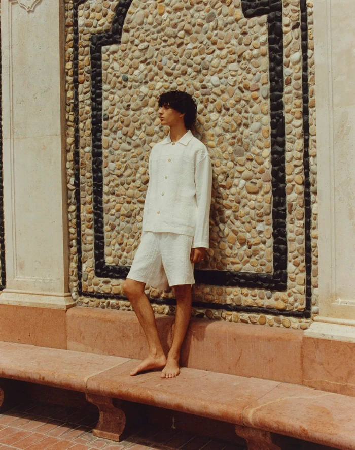 Model Yoesry Detre posing barefoot in a white textured shirt and shorts against a pebble stone wall for COMMAS.