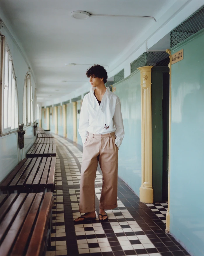 Model Yoesry Detre posing in a white wrap top and wide-leg beige trousers along a curved tiled hallway.