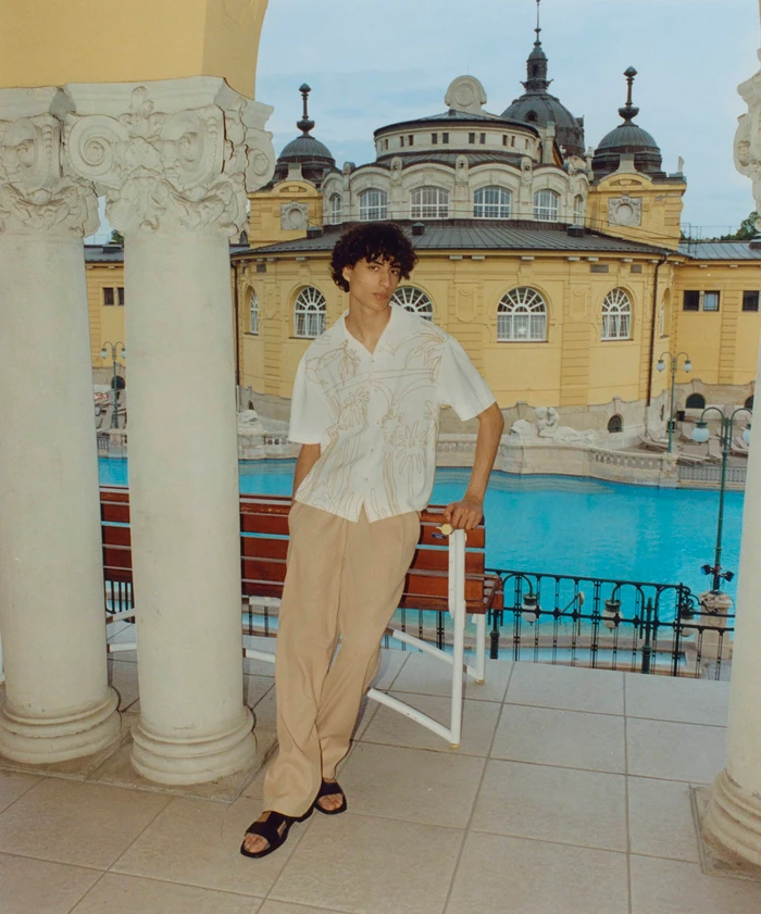 Model Yoesry Detre wearing an embroidered white shirt and beige trousers by a thermal pool in Budapest for COMMAS Resort 2024.