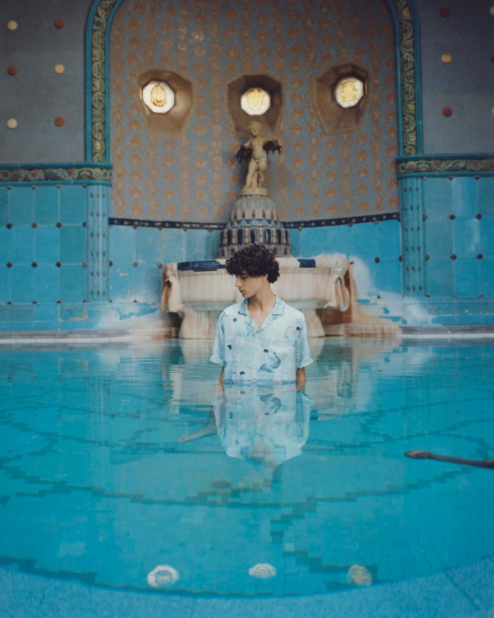 Model Yoesry Detre stands chest-deep in a turquoise thermal pool wearing a pale printed short-sleeve shirt for COMMAS.