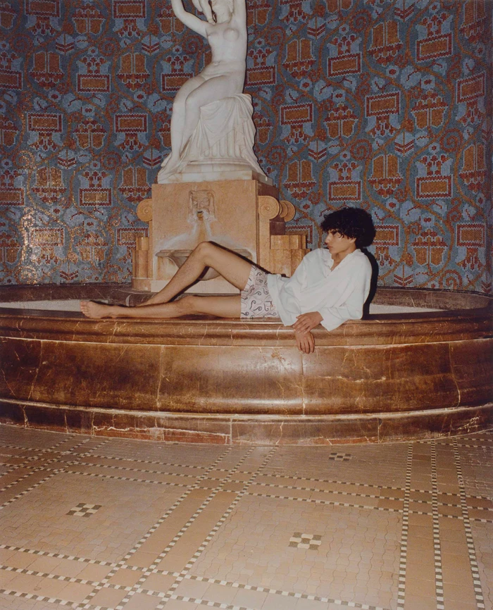 Model lounging on the edge of a marble fountain beneath a white classical sculpture for COMMAS Resort 2024 campaign