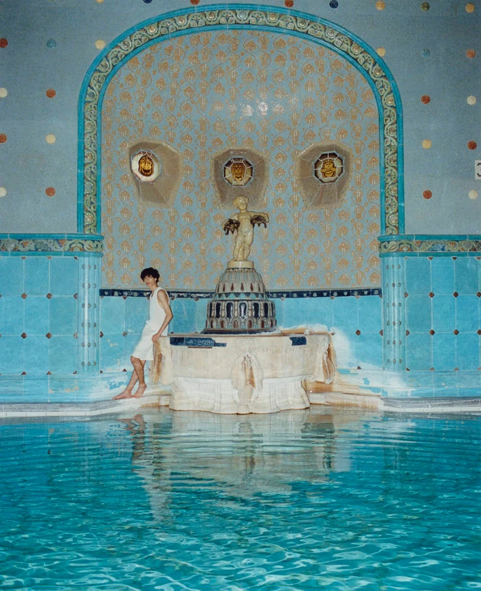 A person in a white top seated next to an ornate tiled fountain beside an indoor swimming pool.