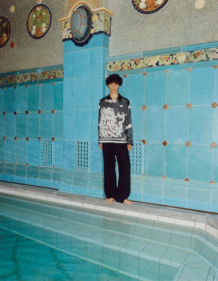 Model standing barefoot by an indoor tiled pool wearing a patterned shirt and dark trousers for COMMAS Resort 2024 campaign