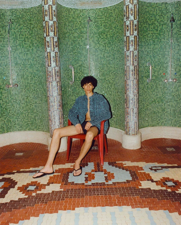 Model Yoesry Detre wearing an unbuttoned blue jacket and swim shorts, seated on a red chair in a mosaic-tiled bathhouse.