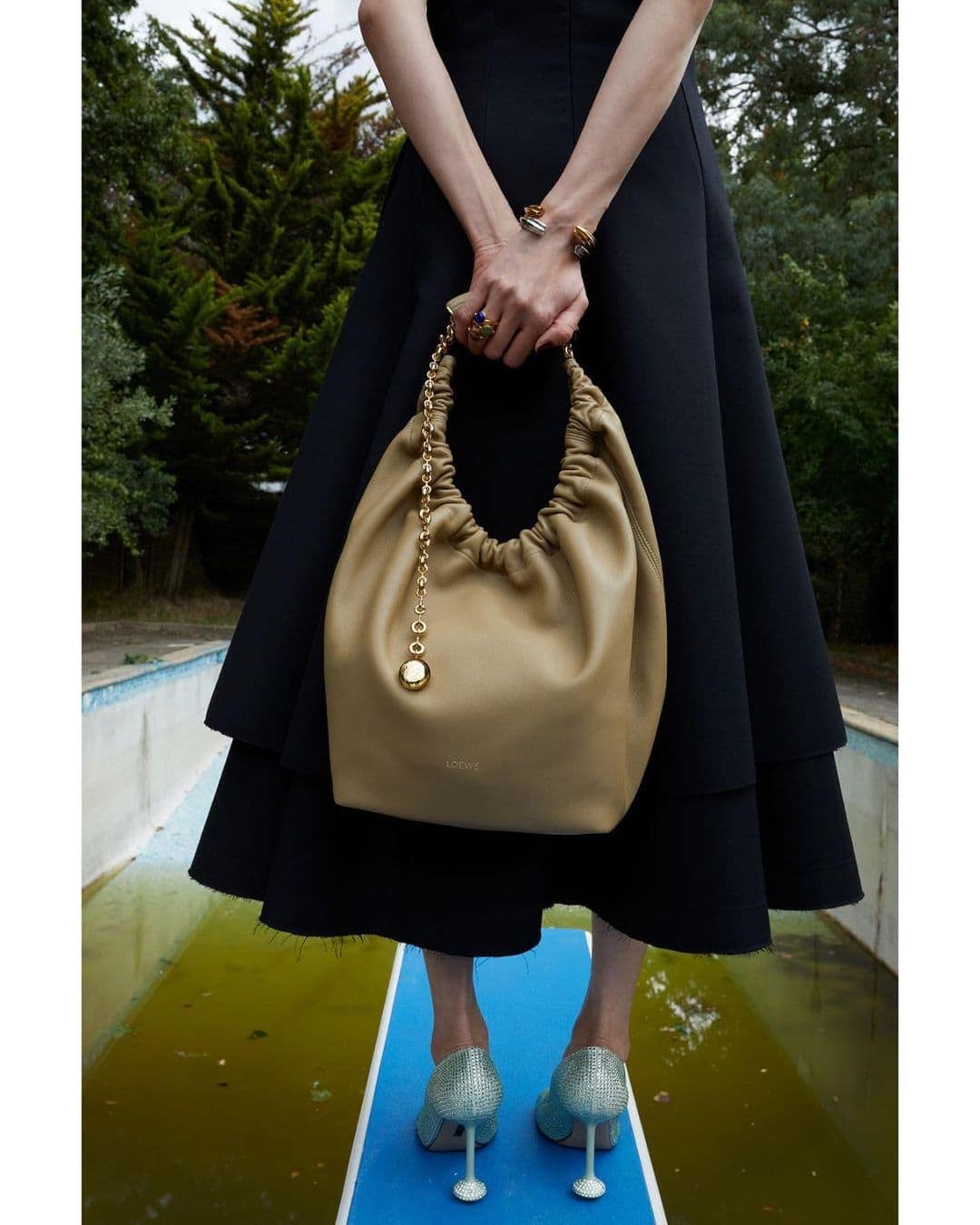 Elegant woman displays a beige leather handbag outdoors. Model wears a black dress and shimmering heels, standing near a pool.
