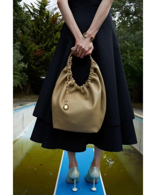 Elegant woman displays a beige leather handbag outdoors. Model wears a black dress and shimmering heels, standing near a pool.