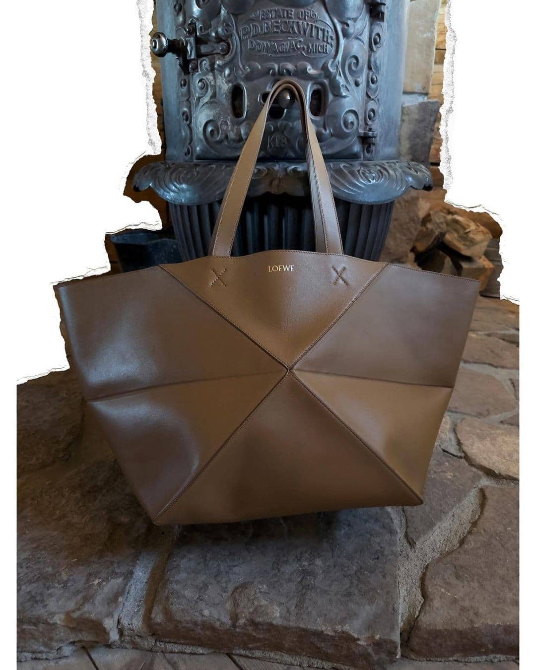 Loewe tan leather origami tote bag with a geometric pattern sits on a stone surface in front of a dark metal antique stove.