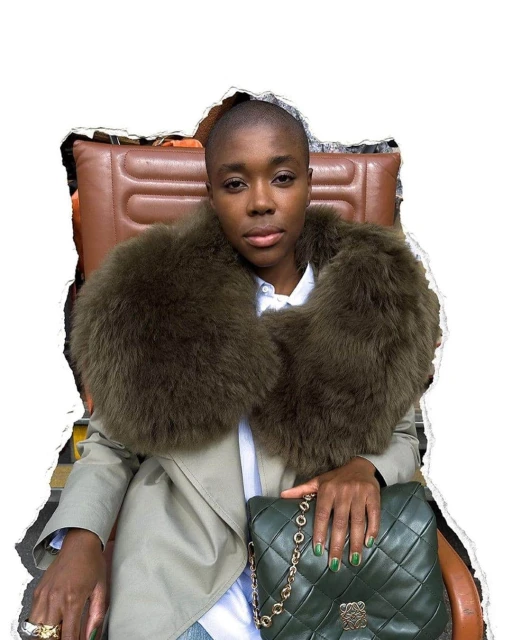 Woman with shaved head wearing a fur stole, trench coat, with a green quilted purse, sitting on an orange leather chair indoors.