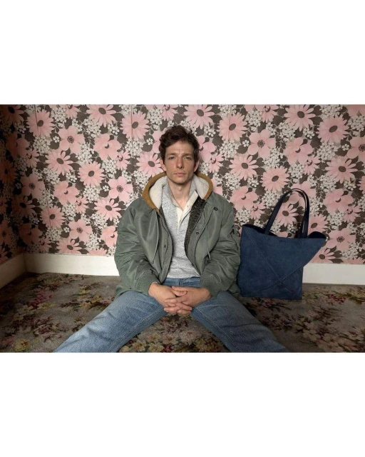 Young man sits indoors against a pink floral wall with a blue tote bag. He wears an olive jacket, grey shirt and jeans in soft light.
