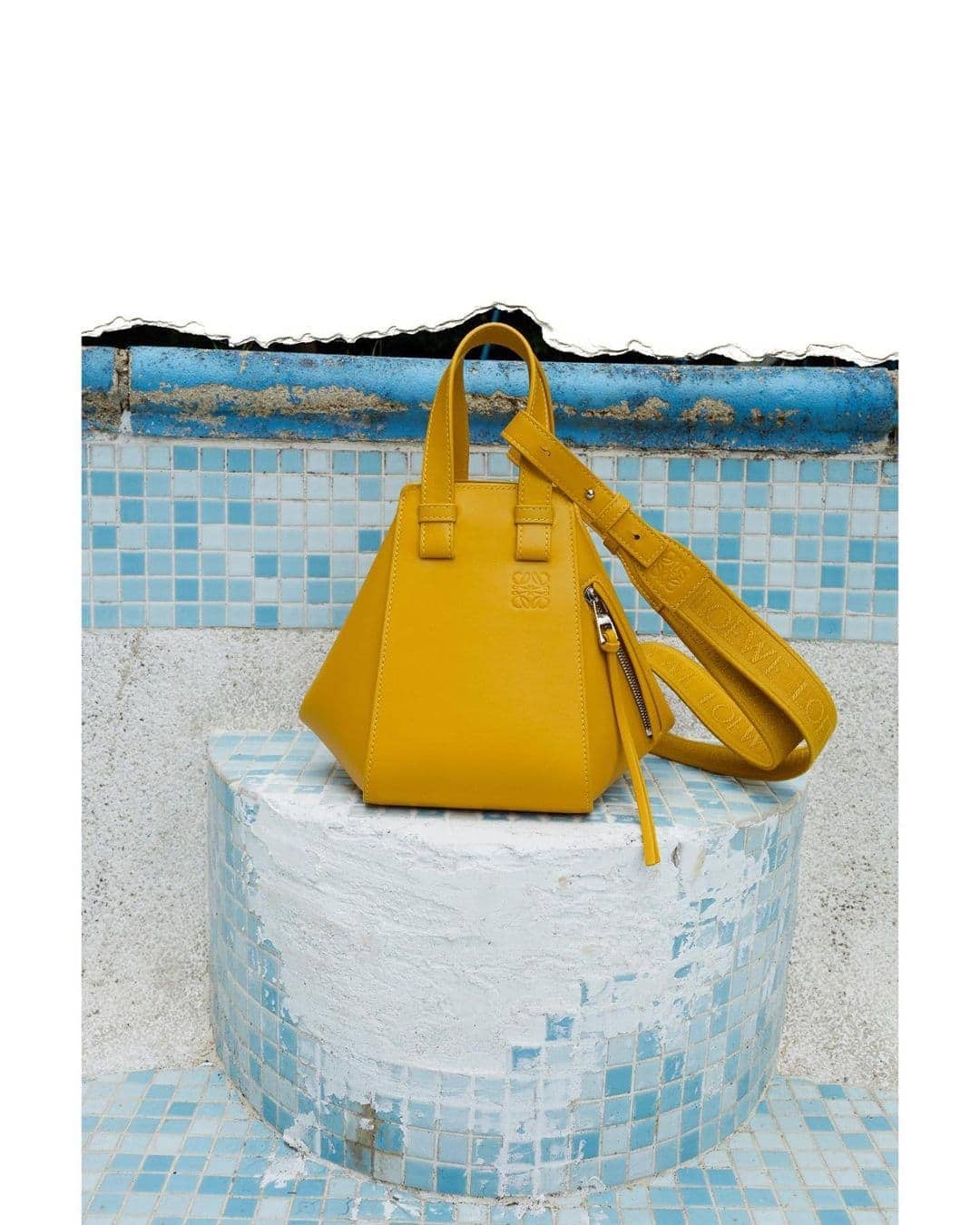 Yellow Loewe bag on a cylindrical base with blue mosaic tile background. Fashion product shot. Luxury handbag with top handles and crossbody strap.
