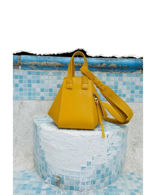 Yellow Loewe bag on a cylindrical base with blue mosaic tile background. Fashion product shot. Luxury handbag with top handles and crossbody strap.