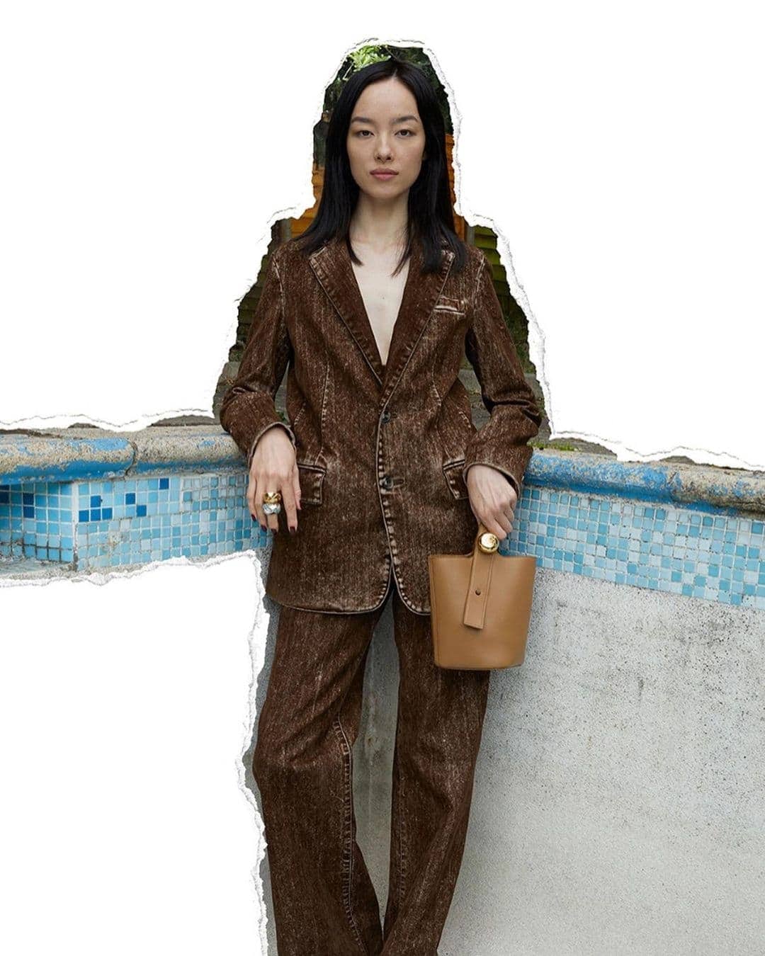 Woman in a brown textured suit poses by a tiled pool with a beige bucket bag and statement rings, set against a white background.