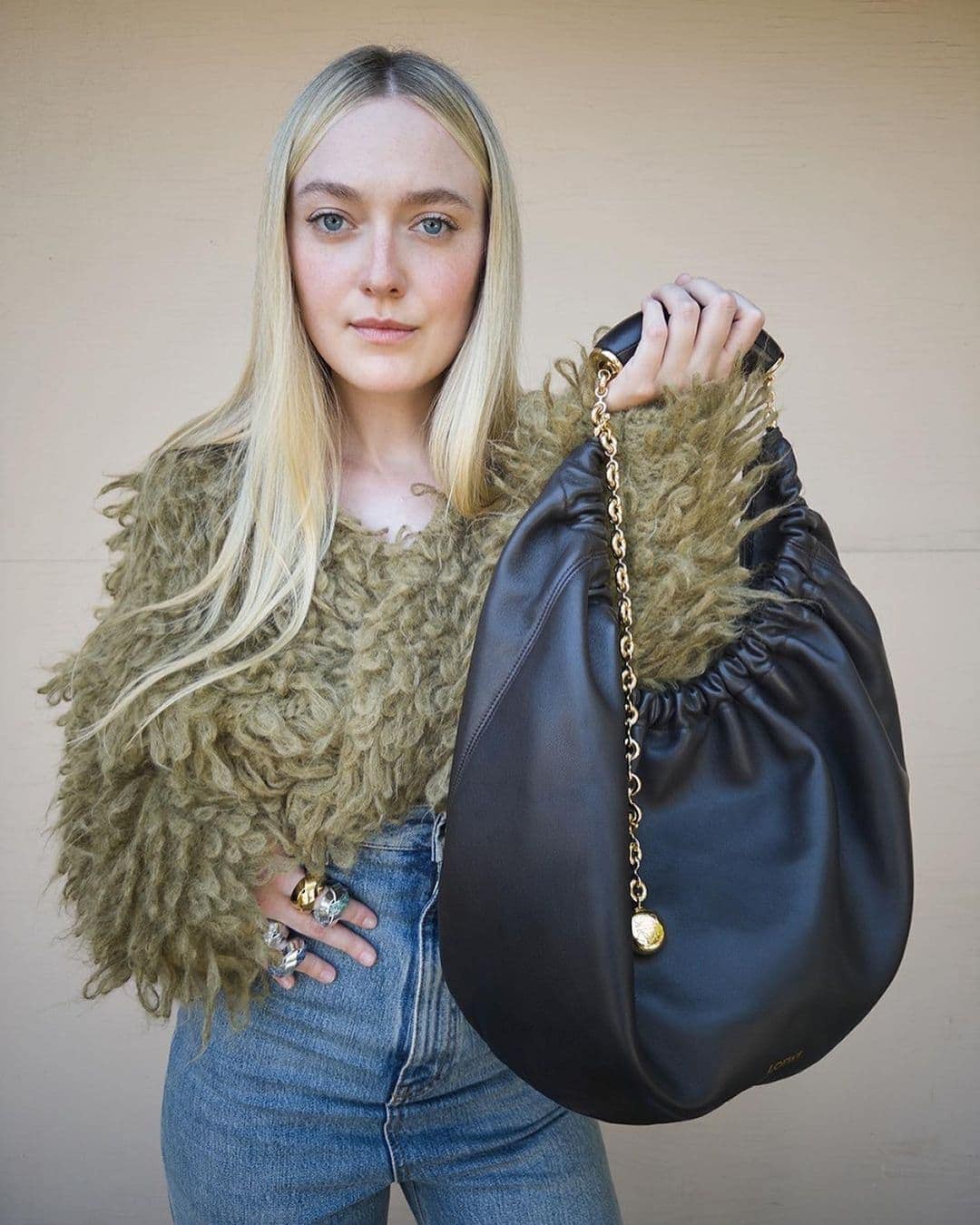 Blonde woman poses with a black leather purse featuring a gold chain strap and wearing a fur-like coat and jeans in a neutral setting.