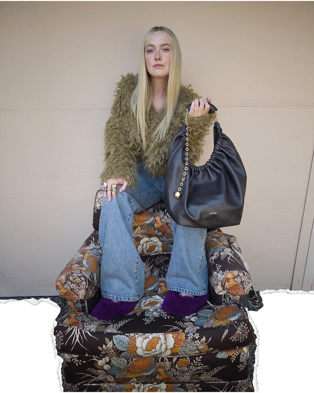 Woman sitting on a floral chair holding a black bag with a chain strap. She wears a furry olive jacket and jeans in an indoor setting.