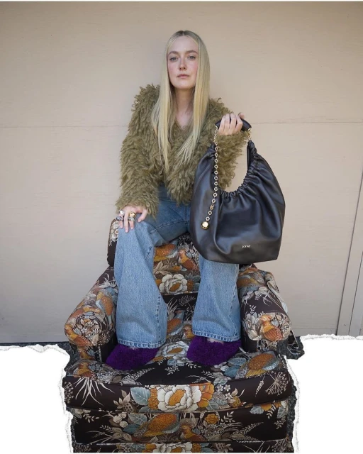 Woman sitting on a floral chair holding a black bag with a chain strap. She wears a furry olive jacket and jeans in an indoor setting.
