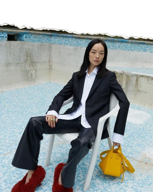 Stylish Asian woman in black blazer and leather pants sitting on a chair with a yellow purse and red slippers in an empty pool.