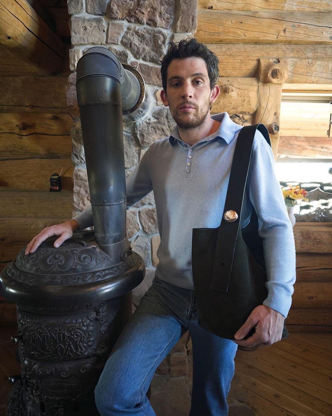 Man in light blue sweater and jeans poses indoors with black leather tote bag and vintage stove against a stone and log wall.