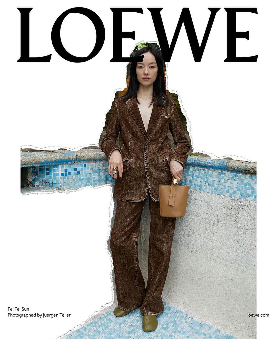 Fashion shot of a young Asian woman in a brown pantsuit with a beige purse standing near a tiled wall and swimming pool with Loewe logo.