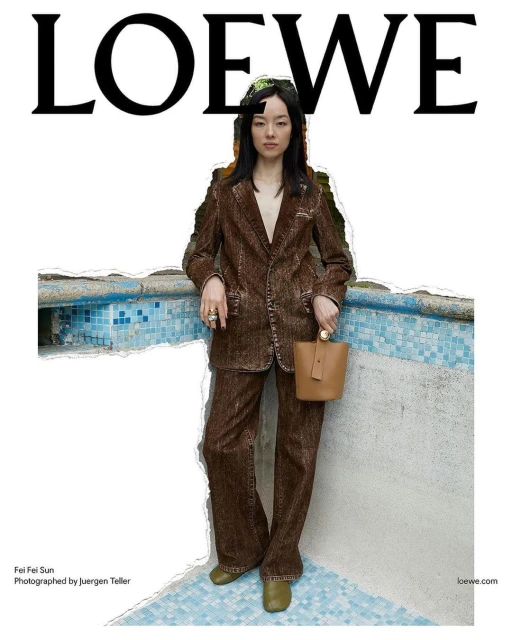 Fashion shot of a young Asian woman in a brown pantsuit with a beige purse standing near a tiled wall and swimming pool with Loewe logo.