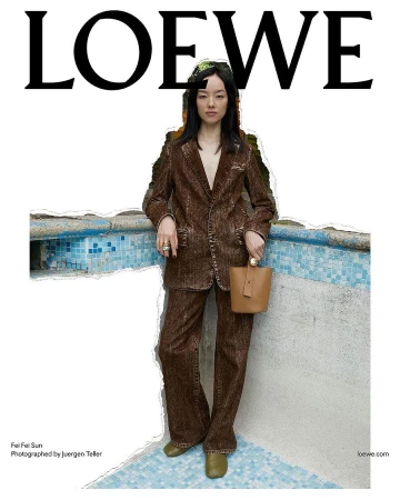 Fashion shot of a young Asian woman in a brown pantsuit with a beige purse standing near a tiled wall and swimming pool with Loewe logo.