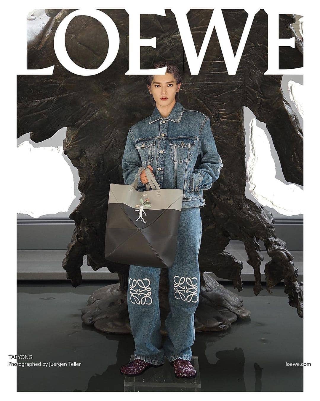 Fashion shot featuring a person in denim jacket and jeans holding a Loewe tote bag, standing against a sculptural backdrop, for a modern, stylish look.