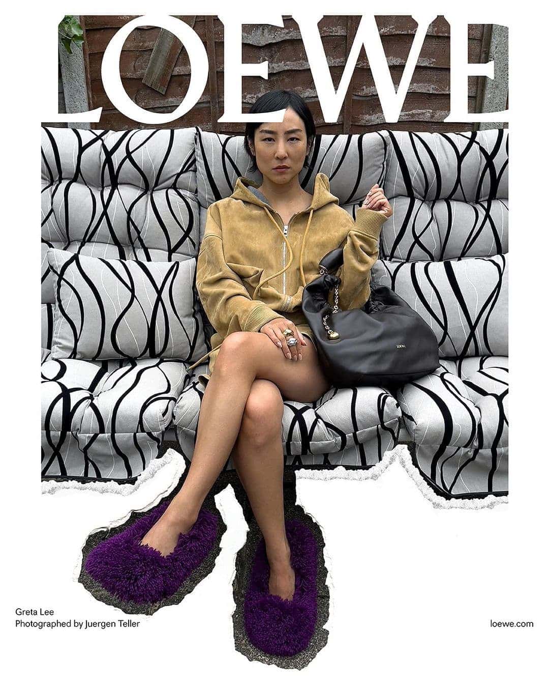 Asian woman poses seated with a black purse, wearing a tan hoodie and large purple fuzzy slippers, in front of a black and white patterned cushion.