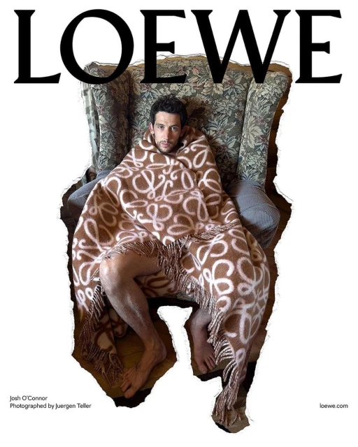 Josh O'Connor sits in a floral armchair covered in a logo-patterned blanket. Shot for Loewe by Juergen Teller in an indoor setting.