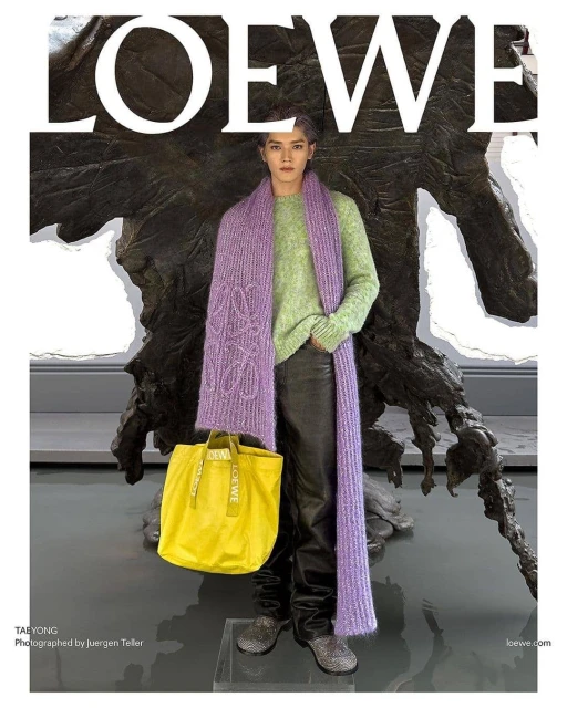 Fashion editorial featuring an adult posing with a yellow tote bag, wearing a green sweater and purple scarf with a stone statue backdrop.