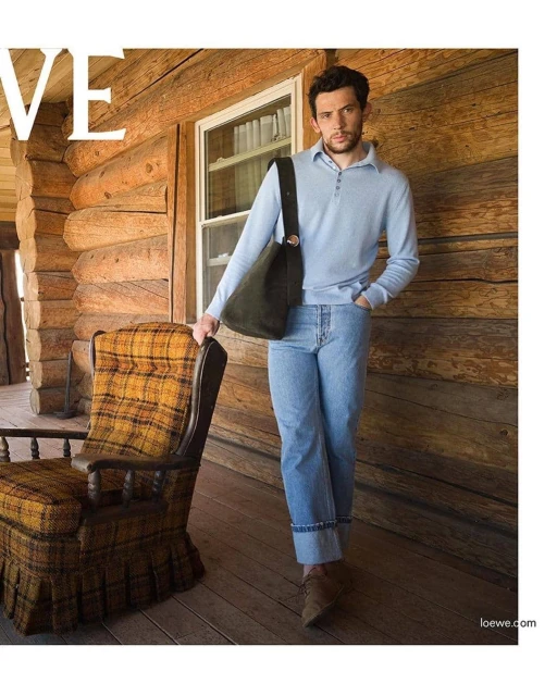 Man in light blue sweater and jeans stands on a wood porch with a brown bag, next to a plaid chair, in front of a log cabin.