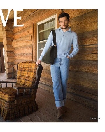 Man in light blue sweater and jeans stands on a wood porch with a brown bag, next to a plaid chair, in front of a log cabin.