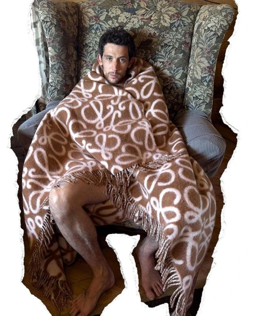 Adult male wrapped in a patterned brown blanket sits in a floral armchair indoors, looking at the camera with a neutral expression.