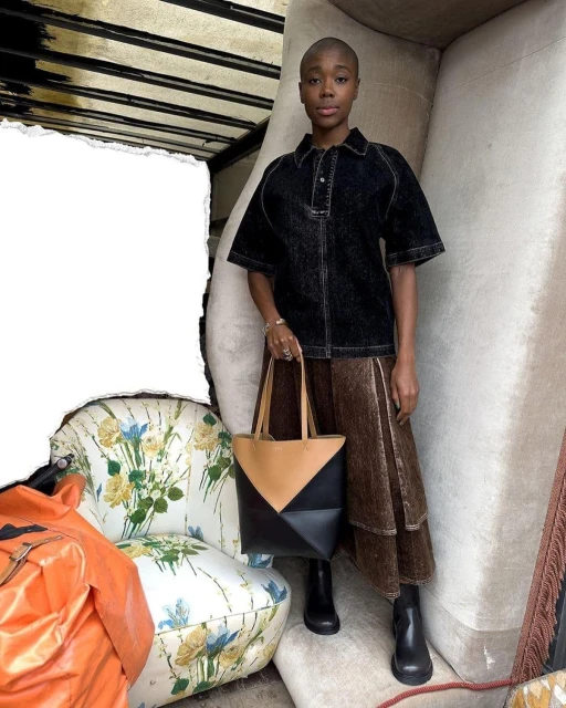 A woman in a black denim shirt and brown skirt stands indoors, holding a geometric tote bag. An armchair is visible.