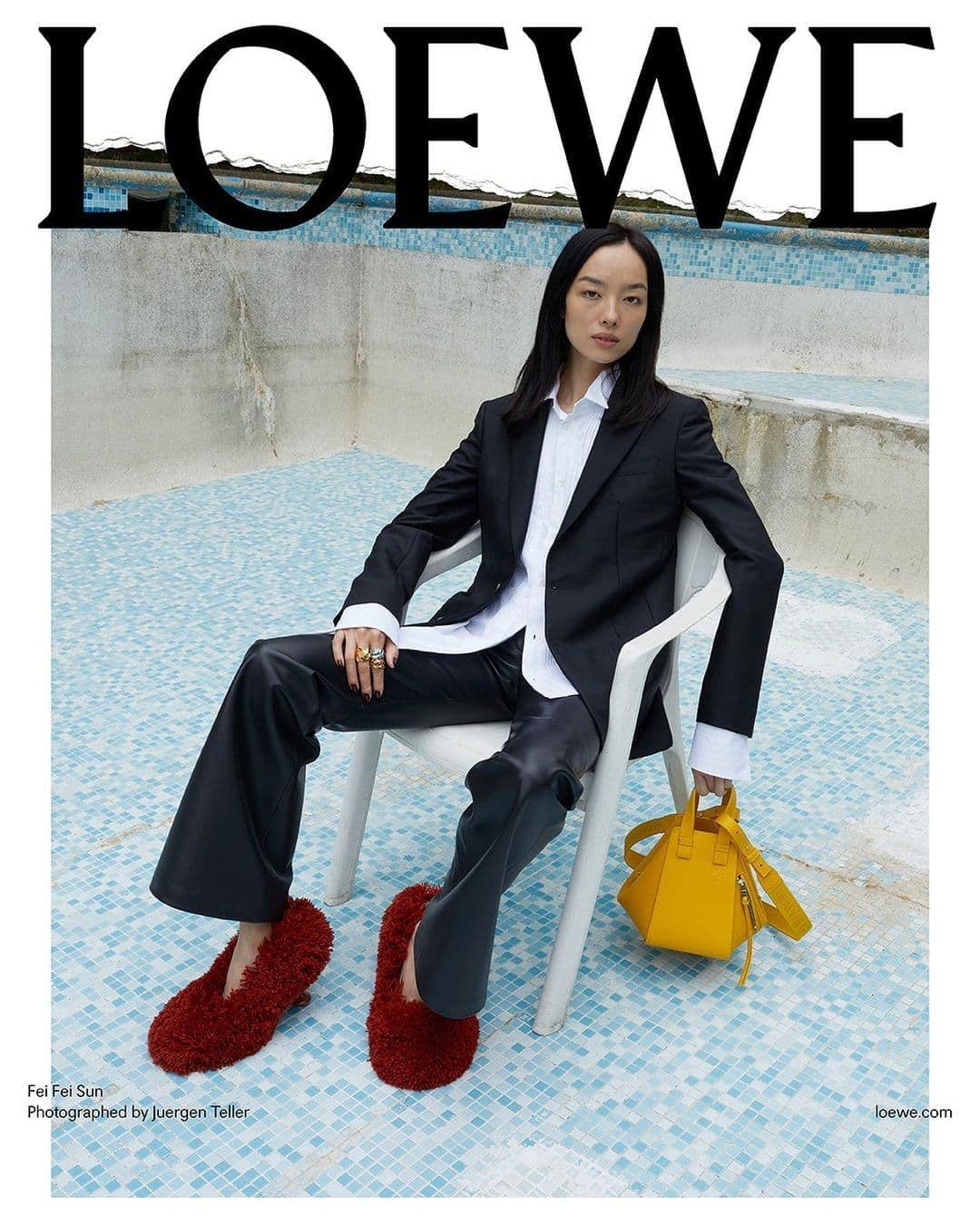 Fashion editorial featuring a woman in a black suit sitting on a white chair with a yellow bag, wearing red slippers, and the Loewe logo.