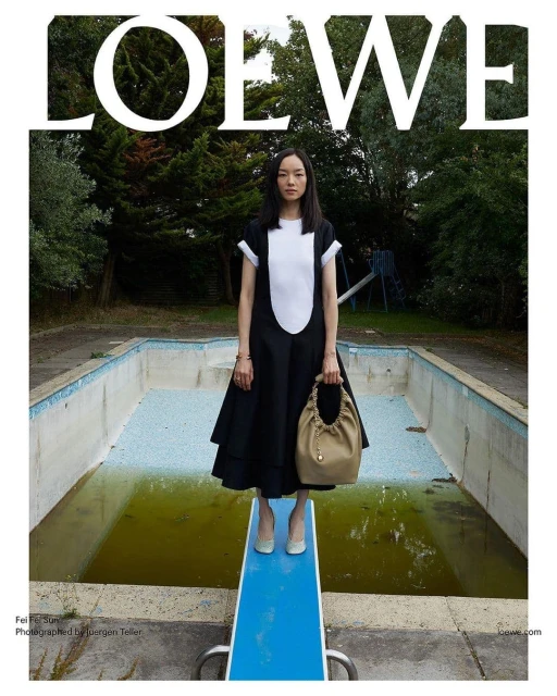 Editorial fashion shot of an Asian woman in a black-and-white dress and beige purse standing on a blue diving board at an abandoned pool.