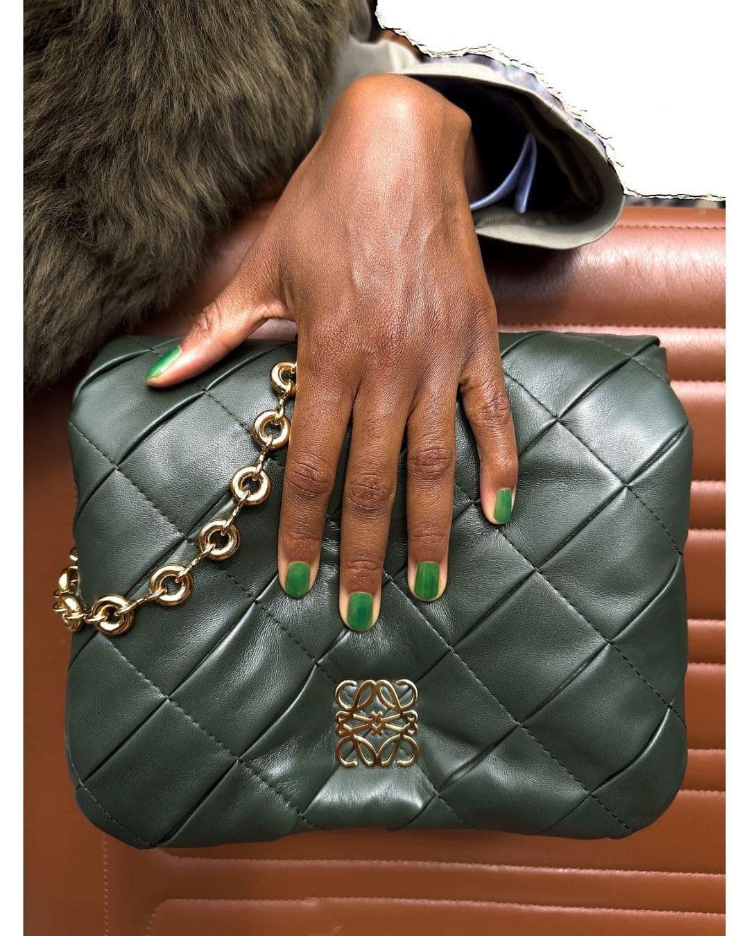 Close-up of a dark-skinned hand with green nail polish holding a quilted green leather purse with a gold chain and logo.