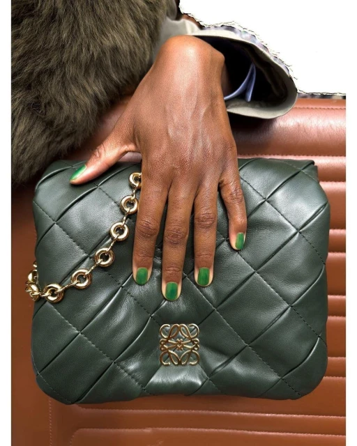 Close-up of a dark-skinned hand with green nail polish holding a quilted green leather purse with a gold chain and logo.