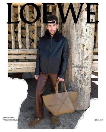 Editorial fashion shot of adult man posing with designer tote bag near woodpile wearing leather jacket and pants for Loewe campaign.