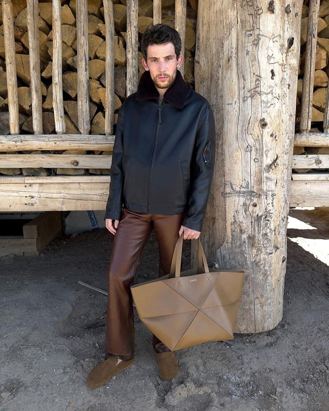 A man in a black jacket, brown pants, and loafers stands near a wood pile, holding a brown tote bag in a rustic outdoor setting.