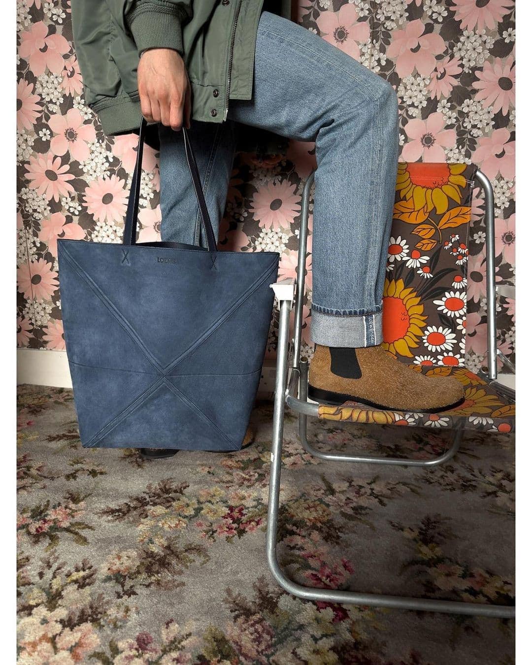Person holding blue tote bag on patterned carpet. The individual sits on a patterned metal folding chair in an indoor setting.