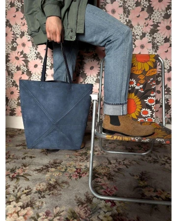 Person holding blue tote bag on patterned carpet. The individual sits on a patterned metal folding chair in an indoor setting.