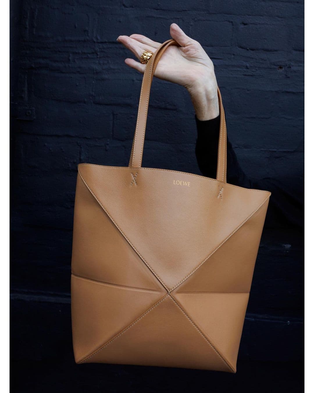 Tan leather Loewe tote bag is held by a hand wearing a gold ring, set against a dark brick wall in a studio setting.