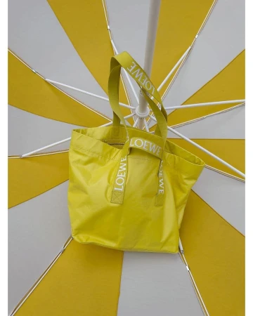 Yellow Loewe tote bag with branded straps hanging against the underside of an open yellow and white striped parasol.