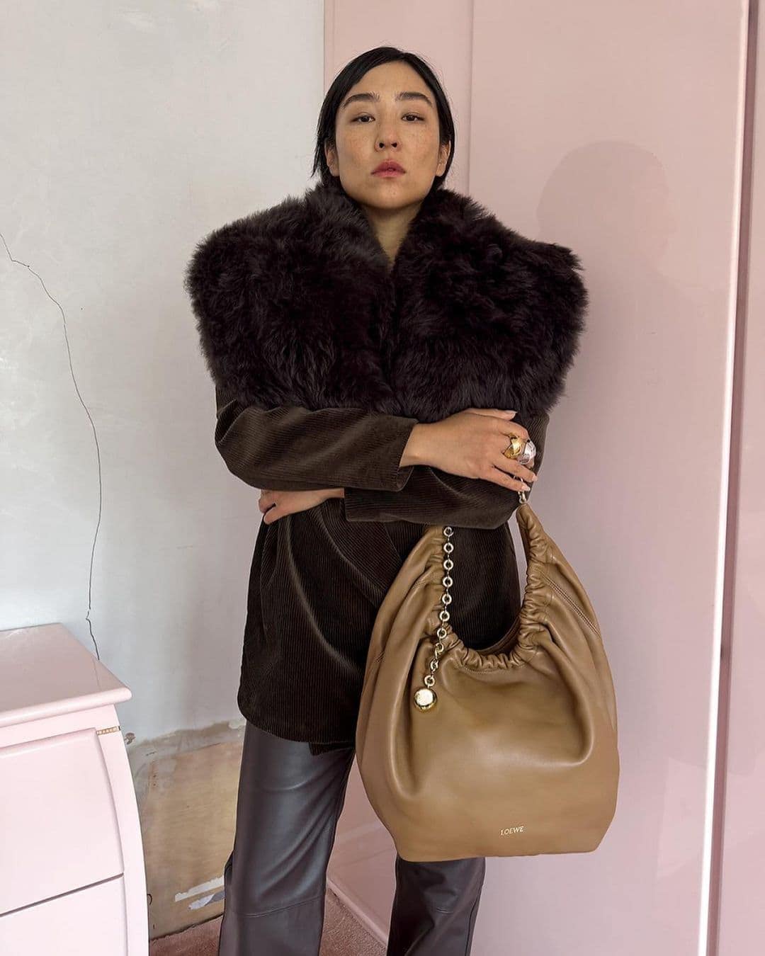A stylish woman wearing a brown jacket with a fur collar and holding a tan purse poses indoors in front of a pink and white wall.