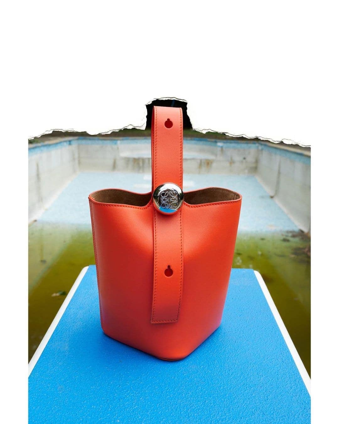 Coral-colored bucket bag with a silver button detail displayed on a blue surface with an out-of-focus outdoor background.