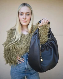 Dakota Fanning holding a large dark leather handbag wearing an olive textured top and blue denim jeans against a plain wall.