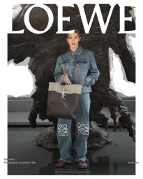 Taeyong wearing a denim jacket and jeans holding a large leather tote bag in front of a dark sculptural background