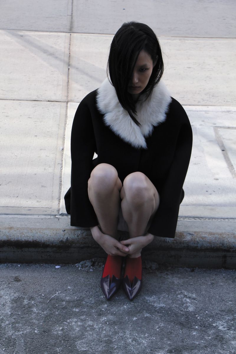 Brunette woman in a black coat with white fur collar sits with knees up. Red and purple high heels. Concrete urban setting.