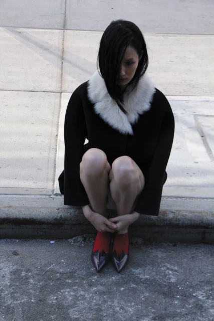 Brunette woman in a black coat with white fur collar sits with knees up. Red and purple high heels. Concrete urban setting.