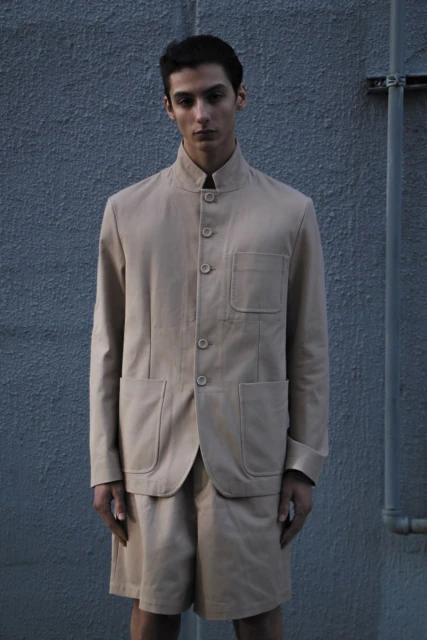 Portrait of a young adult male wearing a beige jacket and shorts posing against a textured gray wall outdoors in neutral lighting.